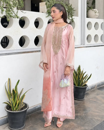 A woman models a light pink traditional outfit with gold embroidery, a matching pink dupatta, and pink sandals, standing in front of a white wall with circular cutouts and potted plants.