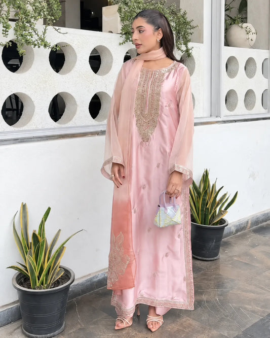 A woman models a light pink traditional outfit with gold embroidery, a matching pink dupatta, and pink sandals, standing in front of a white wall with circular cutouts and potted plants.