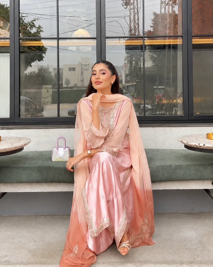 A woman models a light pink traditional outfit with a matching sheer dupatta draped over her shoulders, sitting on a bench in front of a modern building with large black-framed windows.