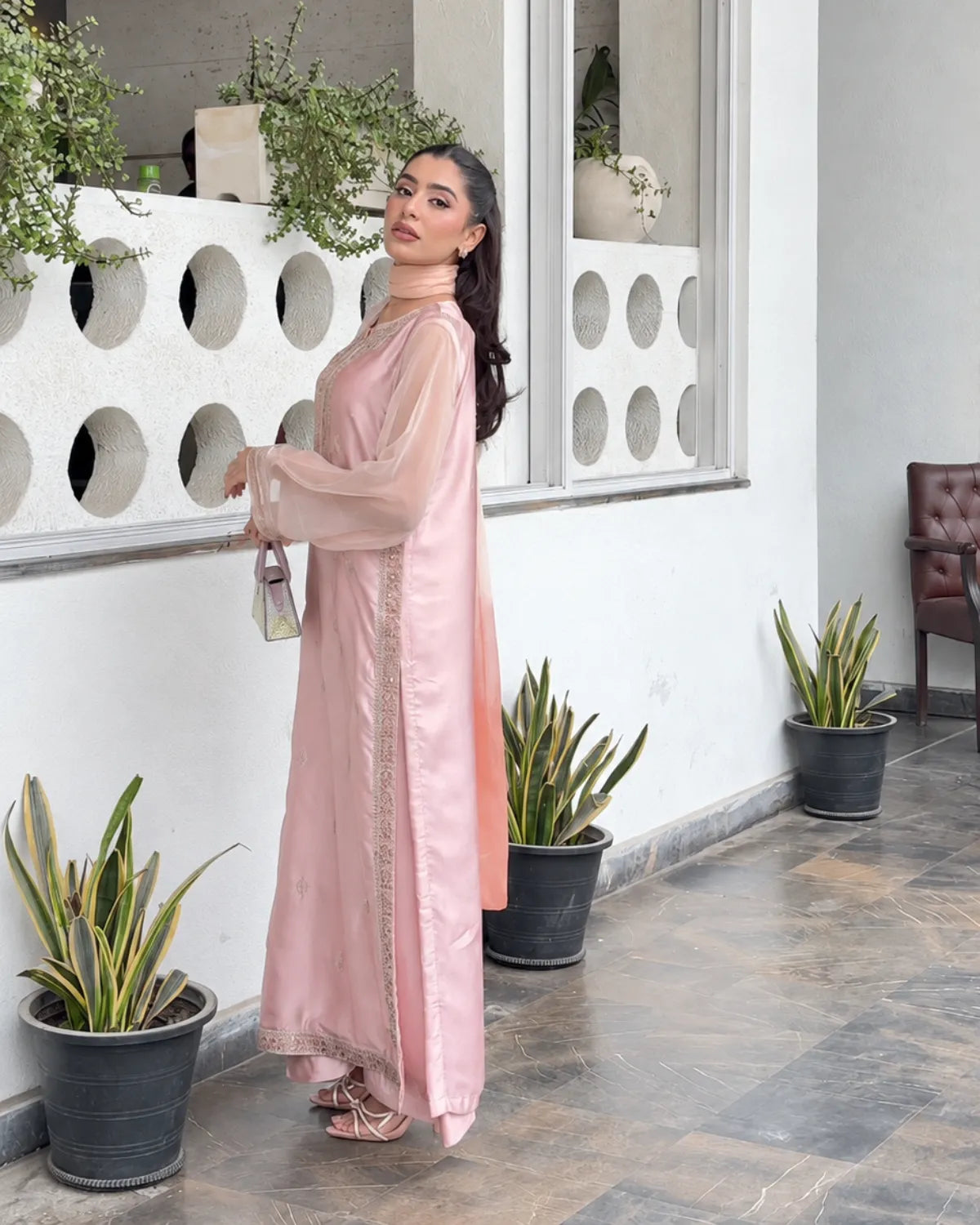 A woman models a light pink traditional outfit with a sheer long-sleeve outer layer and embroidered details, standing in a modern outdoor corridor with potted plants.