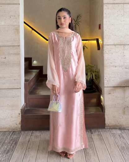 A woman stands in a doorway wearing a light pink traditional long dress with ornate gold embroidery on the chest and hem, paired with a matching sheer scarf and holding a small patterned handbag.