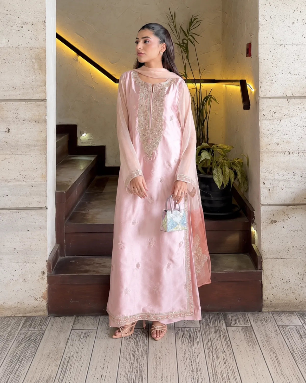 A woman models a light pink traditional outfit with gold embroidery, standing in a doorway with stone walls and wooden stairs behind her.