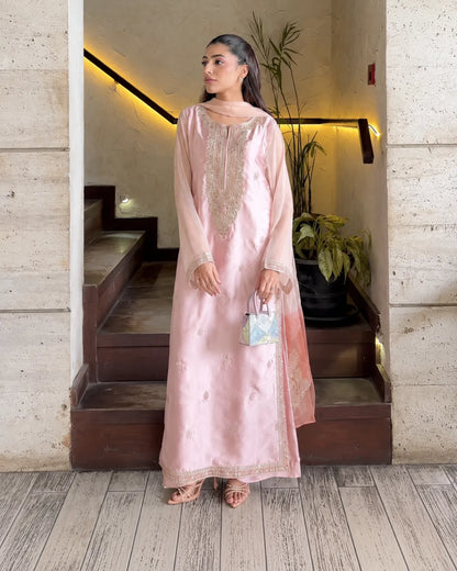 A woman models a light pink traditional outfit with gold embroidery, standing in a doorway with stone walls and wooden stairs behind her.