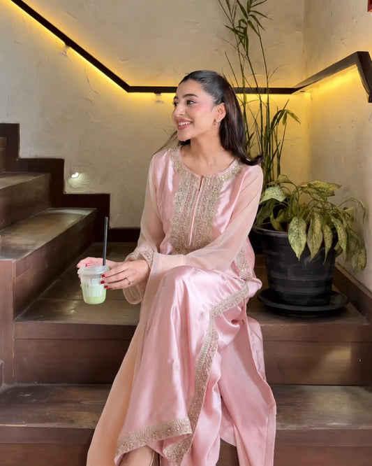 A woman wearing a light pink traditional long dress with gold embroidery sits on wooden stairs in a warm indoor setting, holding a drink.