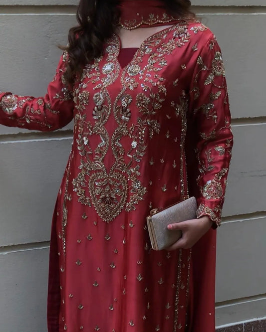 A woman wearing a long-sleeved red embroidered traditional dress with intricate gold metallic embellishments, holding a small metallic clutch against a gray paneled wall.