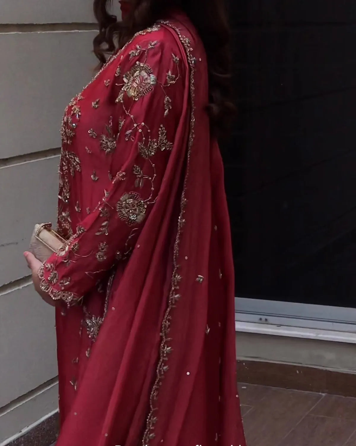 A woman wearing a deep red traditional outfit with intricate gold embroidery and a matching sheer dupatta, holding a small beige clutch.