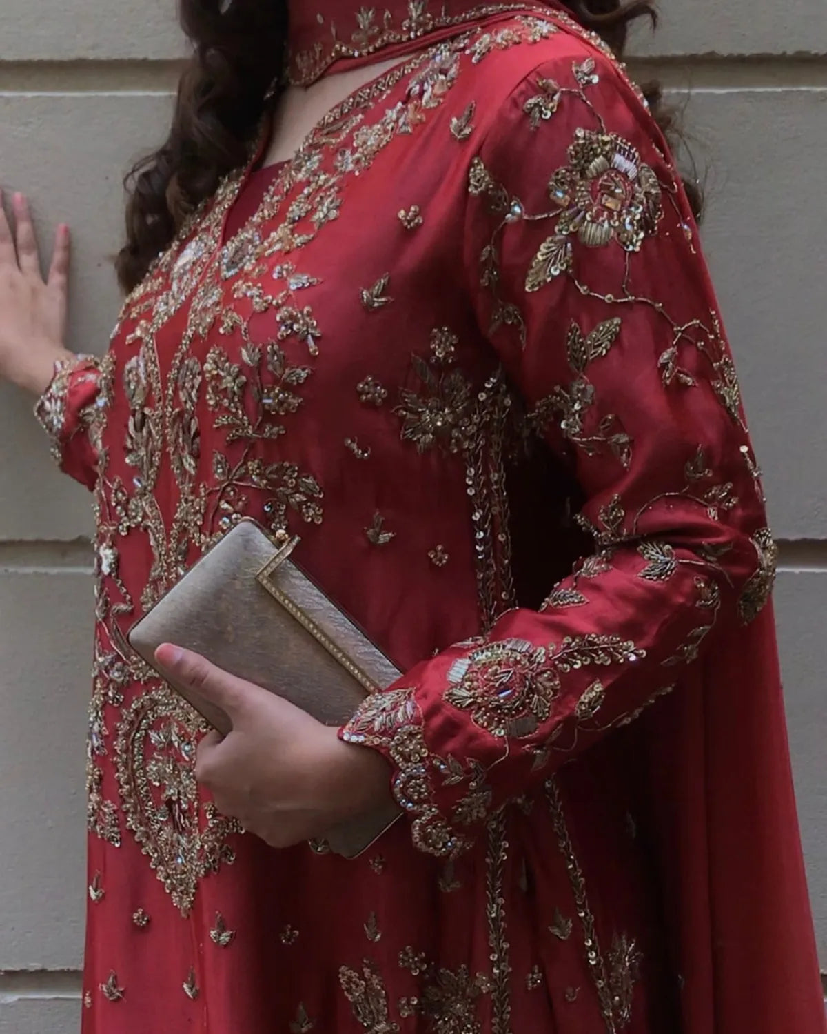 A woman wearing a red embroidered traditional outfit with gold detailing, holding a metallic clutch against a light gray paneled wall.