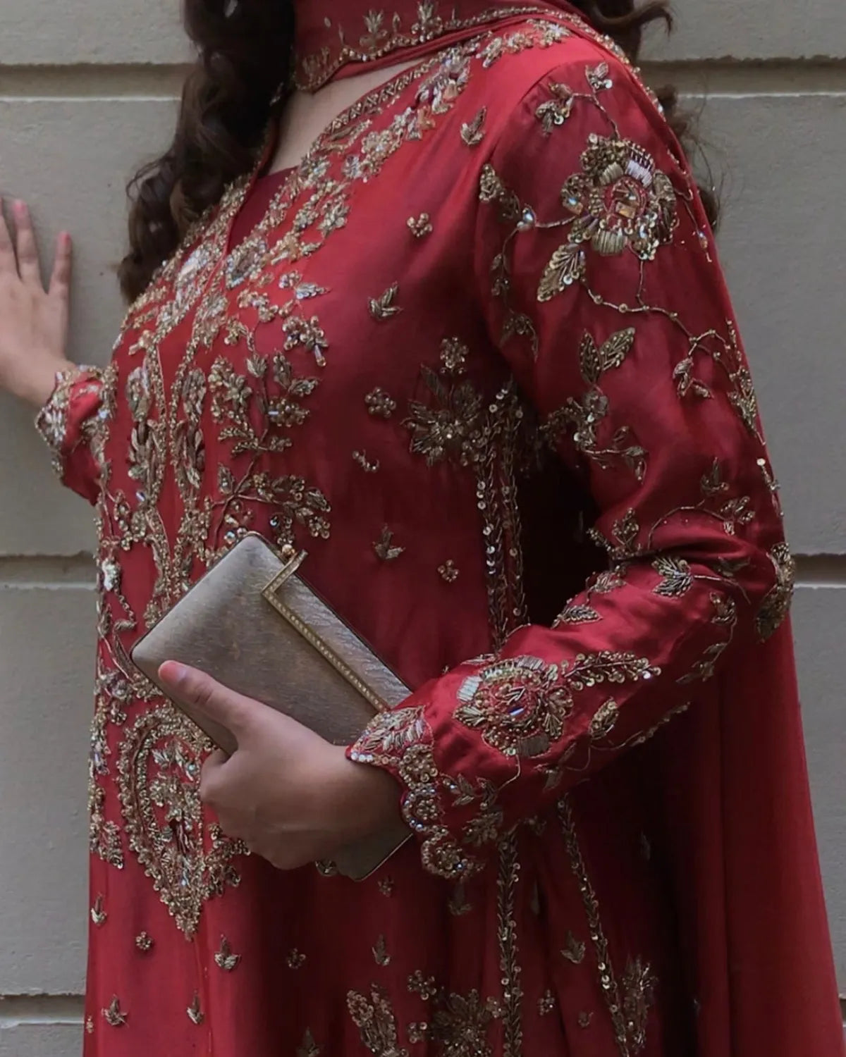 A woman wearing a red embroidered traditional outfit with gold detailing, holding a metallic clutch against a light gray paneled wall.