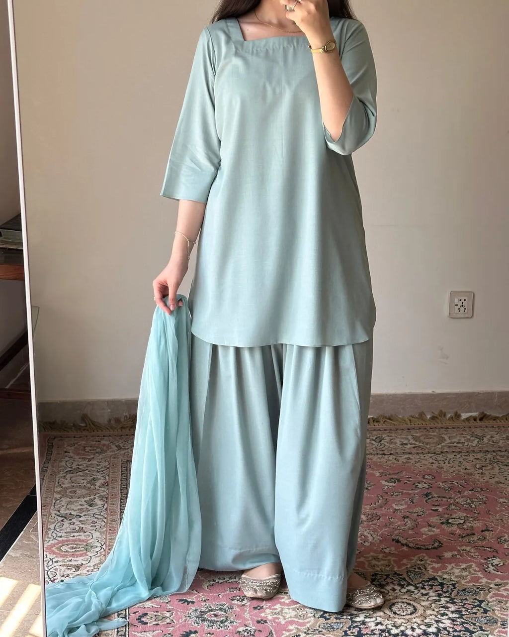 A woman modeling a light blue-green traditional outfit consisting of a square-neck tunic, wide-leg pants, and a matching dupatta, standing in an indoor room with a patterned rug.