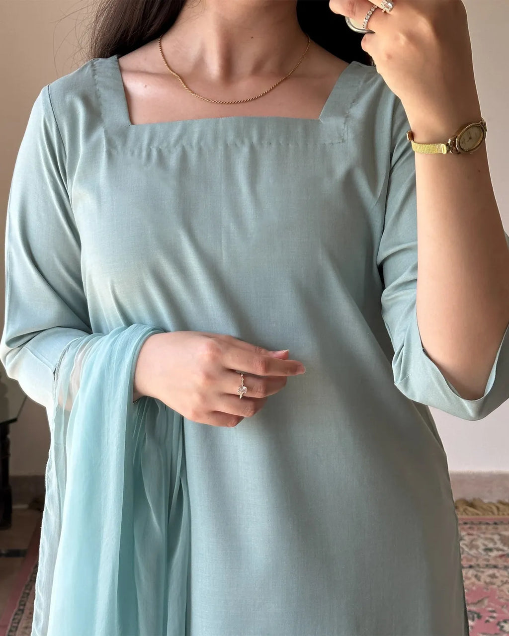 A woman wearing a light sage green or pale mint green traditional tunic dress with a square neckline and a matching sheer dupatta draped over one shoulder. The photo is cropped from the neck down, showing her hands, jewelry, and a watch.