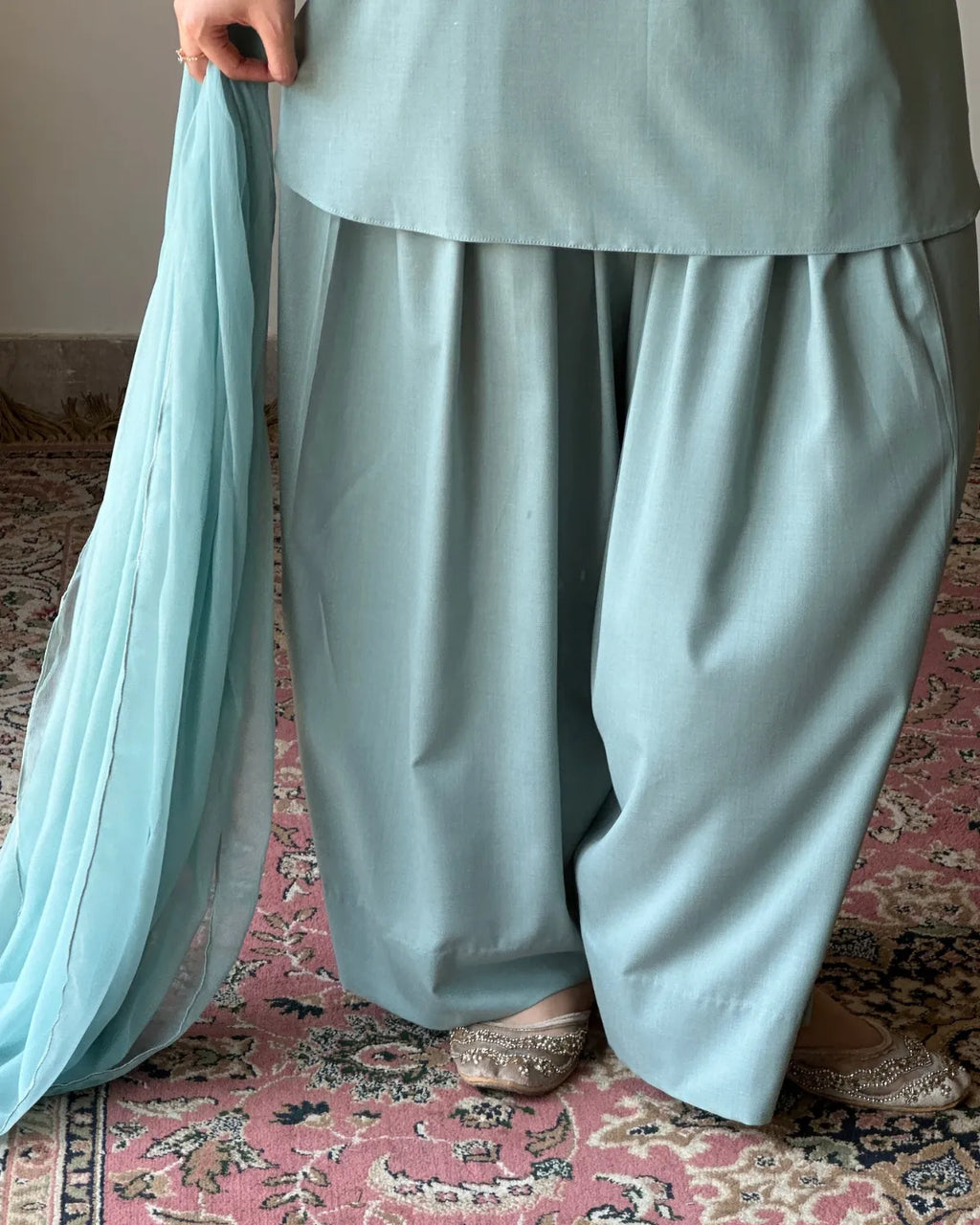 A person wearing a light blue-green traditional outfit with wide-leg pants and a matching sheer dupatta, standing on a patterned pink rug indoors.
