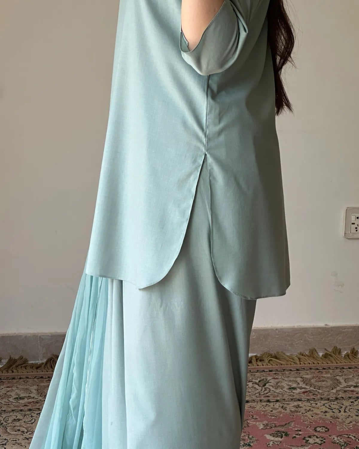 A person wearing a light mint green satin-like tunic top and matching pleated skirt, shown from the back in an indoor setting with a beige wall and patterned rug visible.