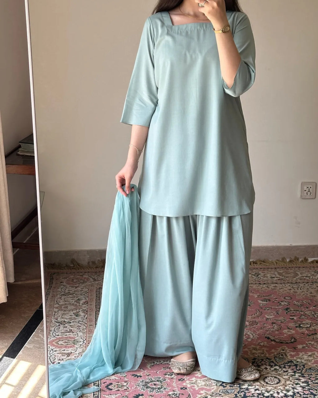 A woman wearing a light blue-green traditional three-piece outfit with a matching dupatta, standing in a room with a patterned rug and mirror.