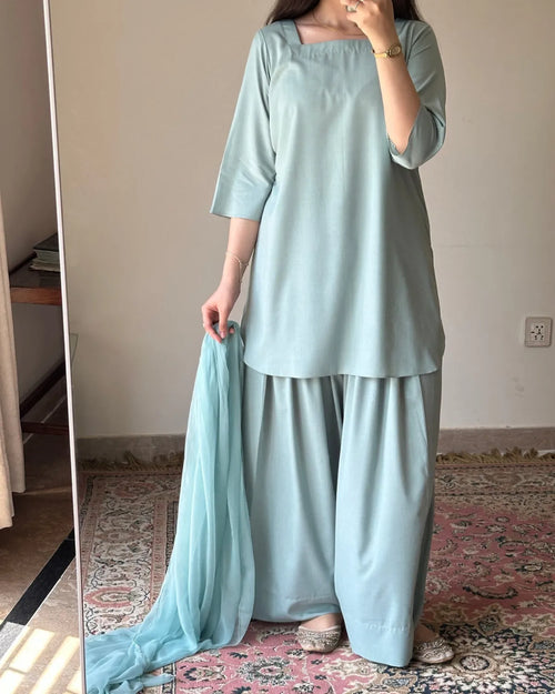 A woman wearing a light blue-green traditional three-piece outfit with a matching dupatta, standing in a room with a patterned rug and mirror.