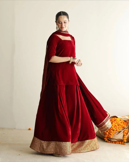 A woman models a traditional deep red velvet sharara suit with a gold zari border, featuring a kurta, flared sharara pants, and a dupatta, set against a plain off-white studio background.