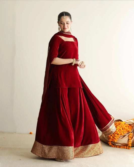 A woman models a traditional deep red velvet sharara suit with a gold zari border, featuring a kurta, flared sharara pants, and a dupatta, set against a plain off-white studio background.