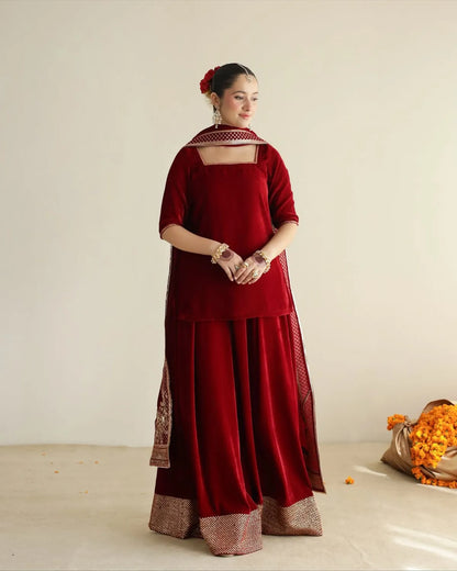 A woman stands in a studio wearing a deep red traditional outfit with a matching dupatta and ornate metallic border details. She is photographed against a plain light beige wall with a small bouquet of orange flowers on the floor to the right.