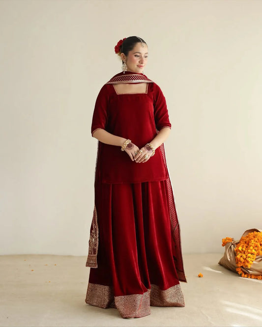 A woman stands in a studio wearing a deep red traditional outfit with a matching dupatta and ornate metallic border details. She is photographed against a plain light beige wall with a small bouquet of orange flowers on the floor to the right.