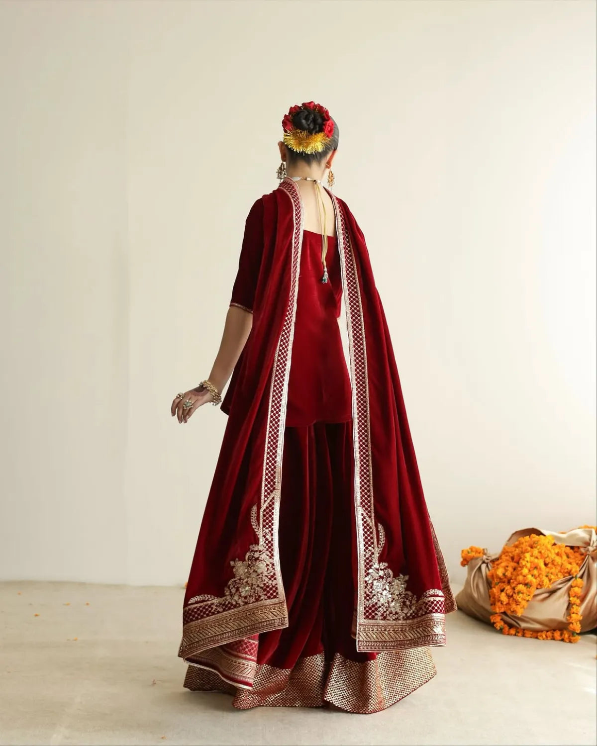 A woman stands with her back to the camera wearing a deep red velvet traditional outfit with ornate gold embroidery and a matching draped outer layer. The setting is a minimalist cream-colored studio with a small decorative garland on the floor.