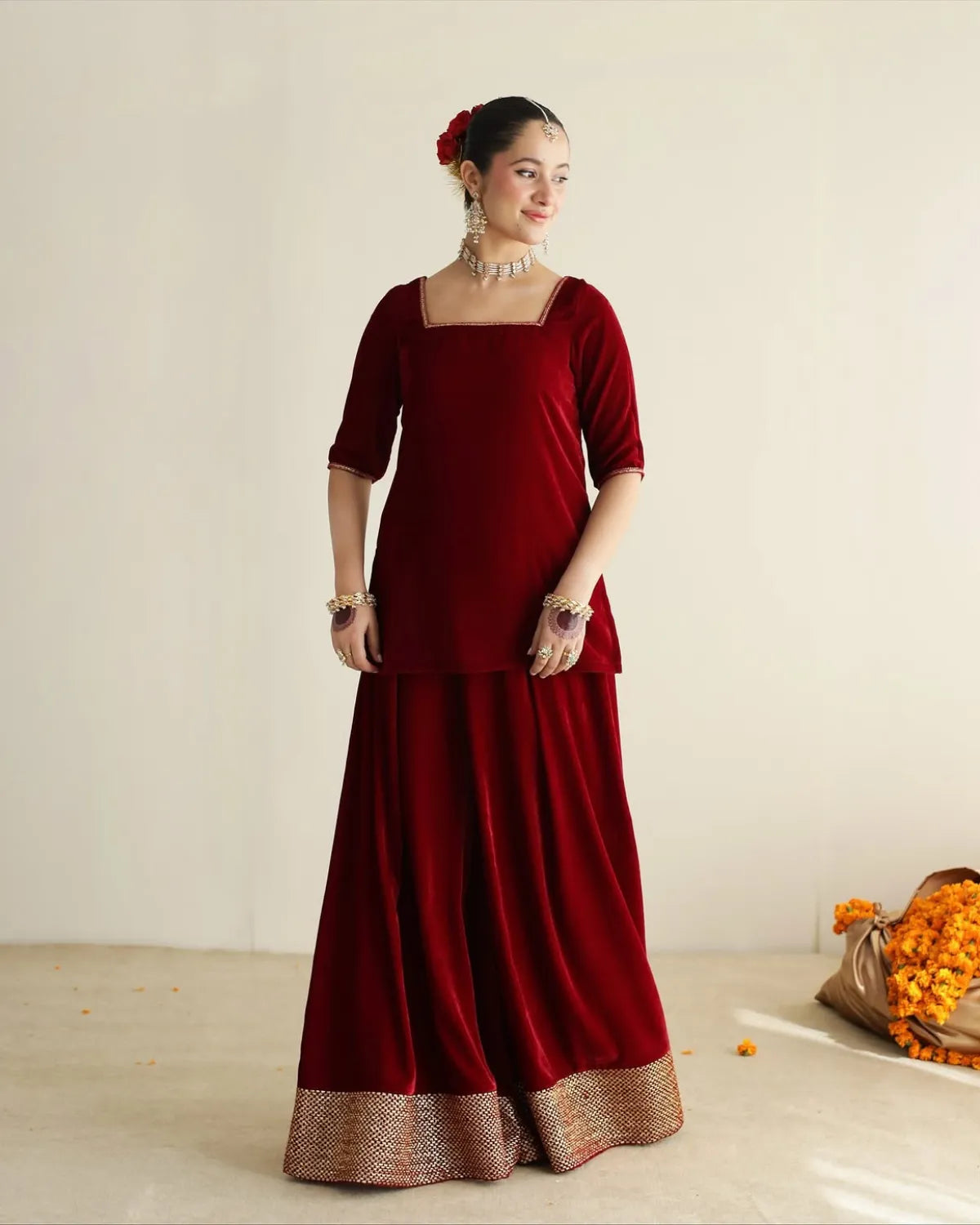 A woman models a deep maroon velvet traditional outfit consisting of a square-neck tunic with elbow-length sleeves and a matching wide flared skirt. The skirt features a decorative metallic gold border at the hem.