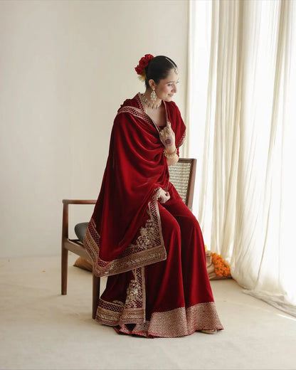 A woman seated on a wooden chair in a bright indoor setting, wearing a deep red traditional outfit with ornate gold embroidery and a matching draped shawl. She has a red flower in her hair and is posed beside sheer white curtains.