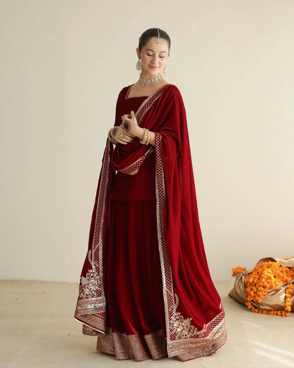 A woman models a deep maroon velvet traditional outfit with ornate gold embroidery and a matching draped dupatta, set against a plain beige studio background.