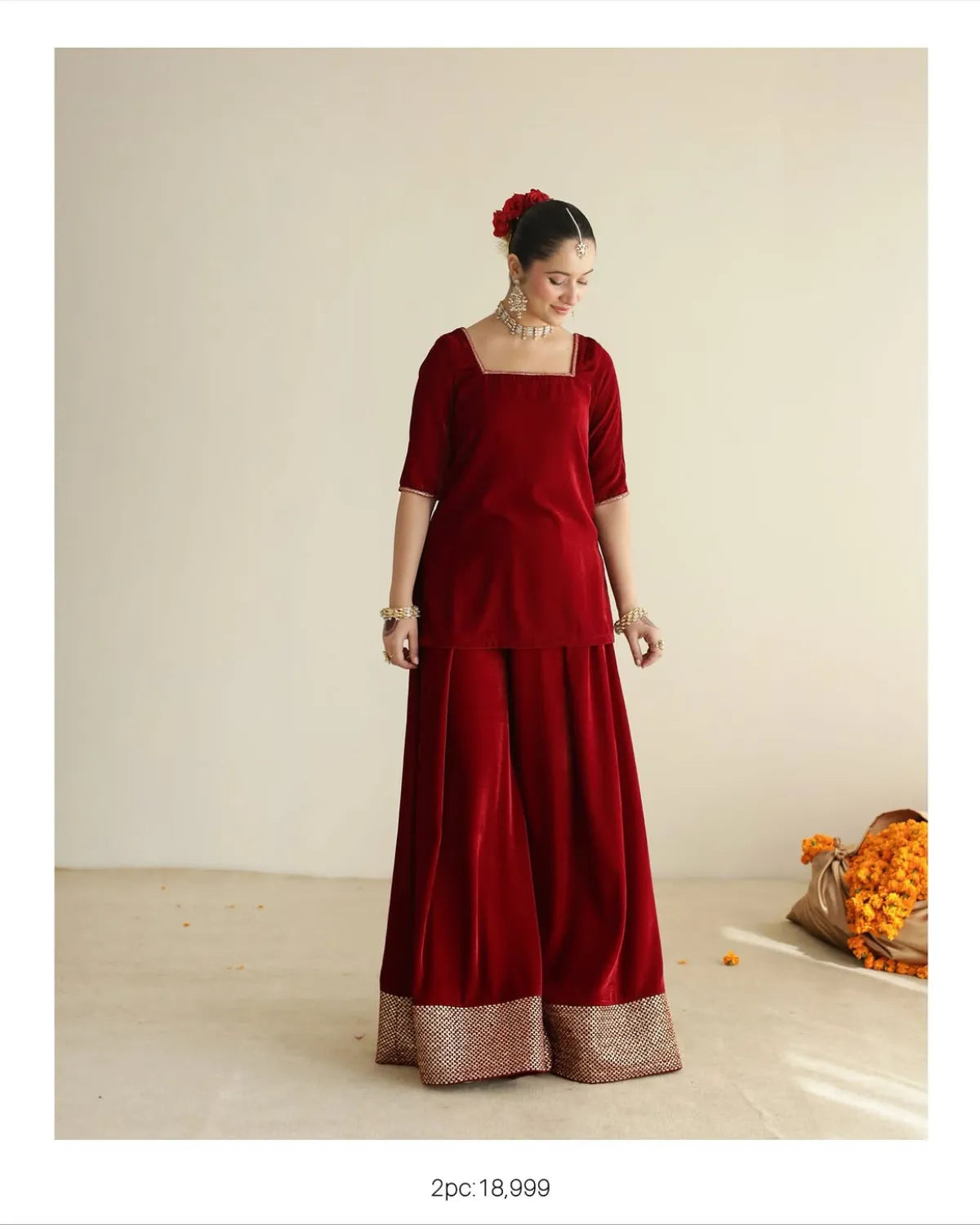 A woman models a deep red velvet traditional outfit consisting of a square-neck top with short sleeves and wide-leg pants with a metallic gold border. She is standing in a studio with a beige background and a bag of orange flower petals on the floor.