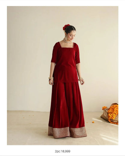 A woman models a deep red velvet traditional outfit consisting of a square-neck top with short sleeves and wide-leg pants with a metallic gold border. She is standing in a studio with a beige background and a bag of orange flower petals on the floor.