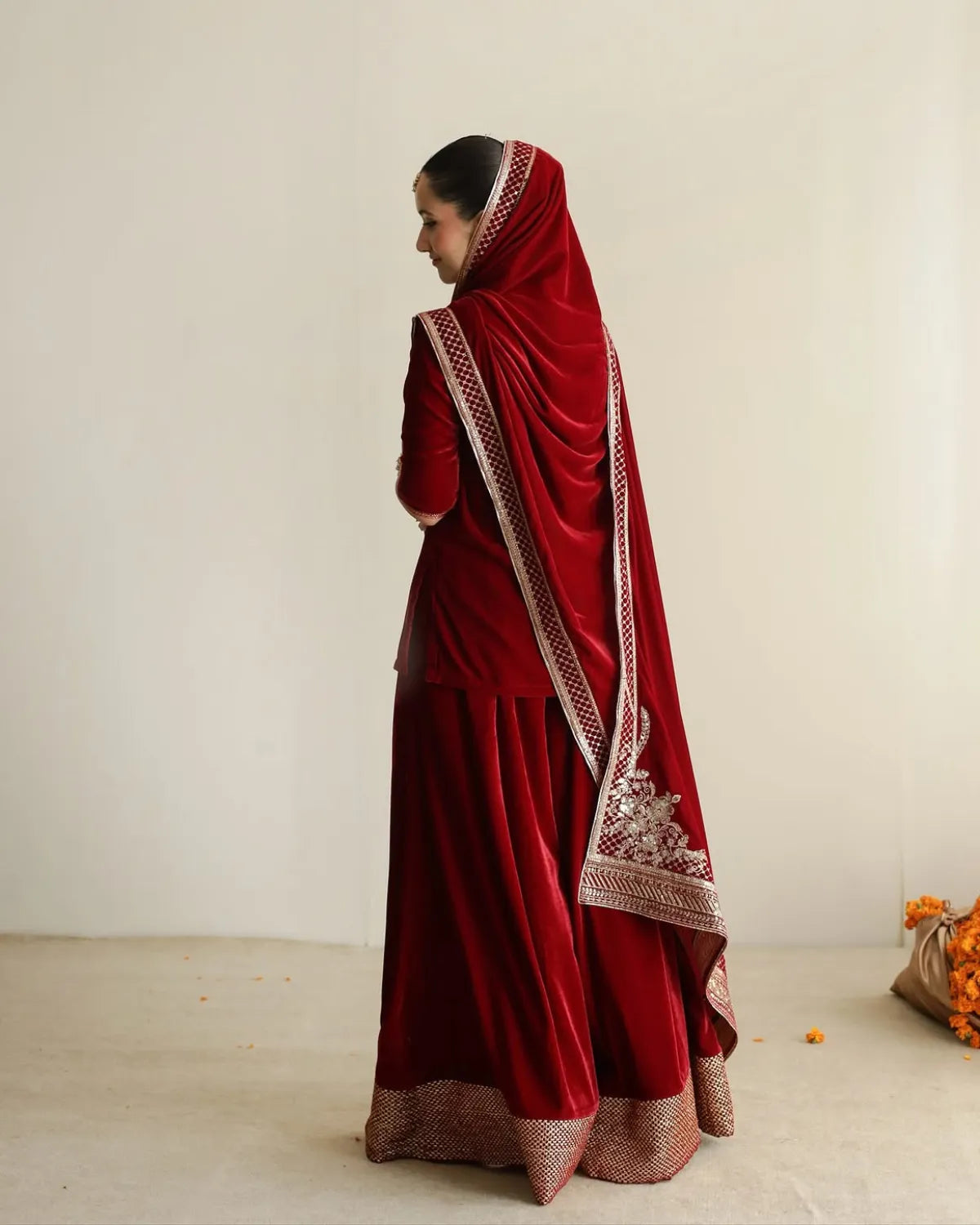 A woman wearing a traditional deep red velvet salwar suit with intricate gold embroidery and a matching dupatta, shown from a side-back angle against a plain off-white studio background.