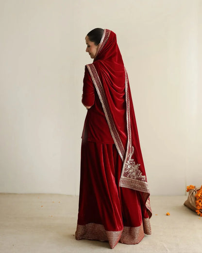 A woman wearing a traditional deep red velvet salwar suit with intricate gold embroidery and a matching dupatta, shown from a side-back angle against a plain off-white studio background.