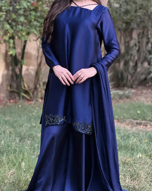 A woman wearing a long navy blue satin dress with floral embroidered trim near the hem, standing outdoors on grass with trees in the background.