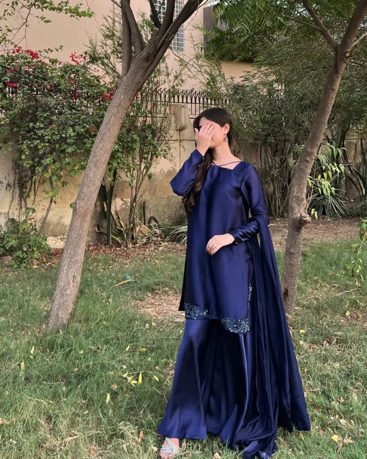 A woman wearing a navy blue traditional outfit with a matching draped scarf, standing outdoors in a grassy garden area with trees and a building in the background.