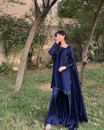 A woman wearing a navy blue traditional outfit with a matching draped scarf, standing outdoors in a grassy garden area with trees and a building in the background.