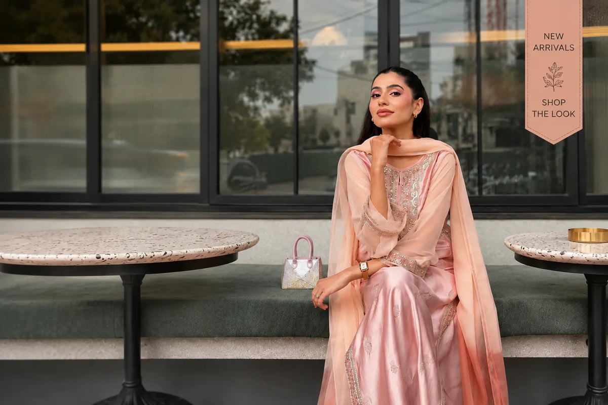 A woman models a light pink traditional outfit with a matching sheer dupatta, sitting on a green bench between two stone tables in front of a modern glass building. A vertical banner on the right reads 'NEW ARRIVALS SHOP THE LOOK'.